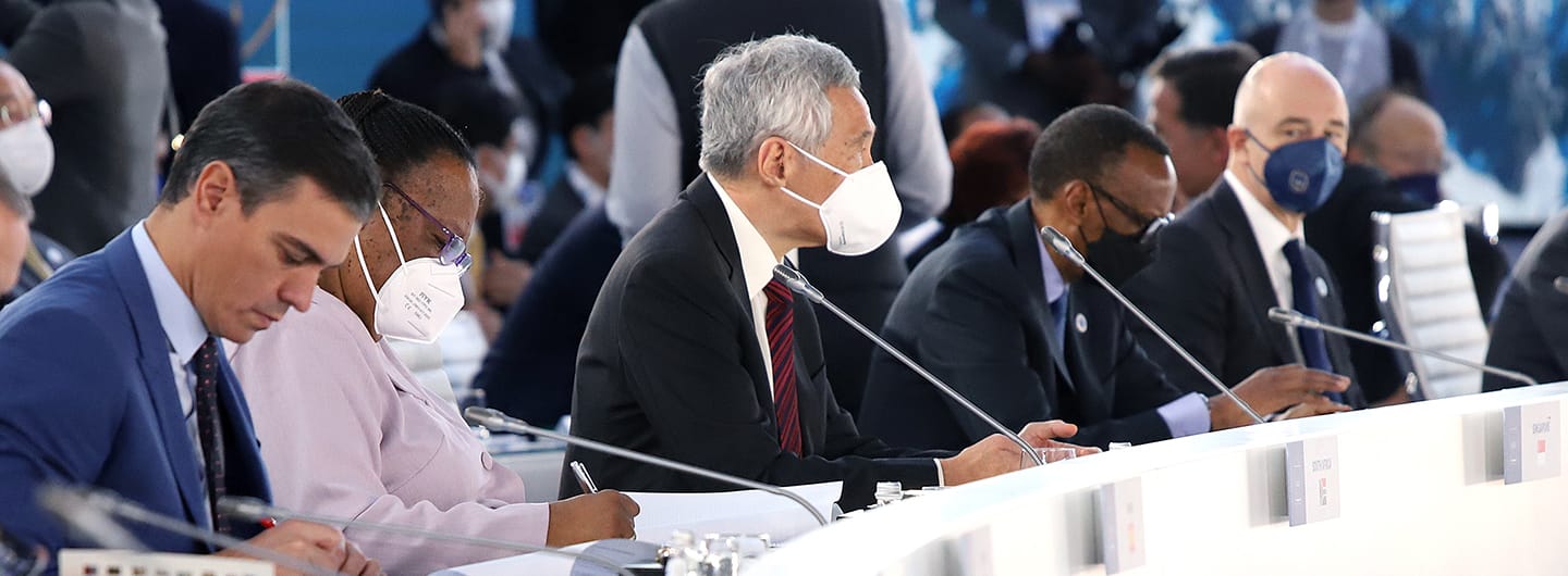 Masked officials at long table, attending a formal international meeting. Microphones present.